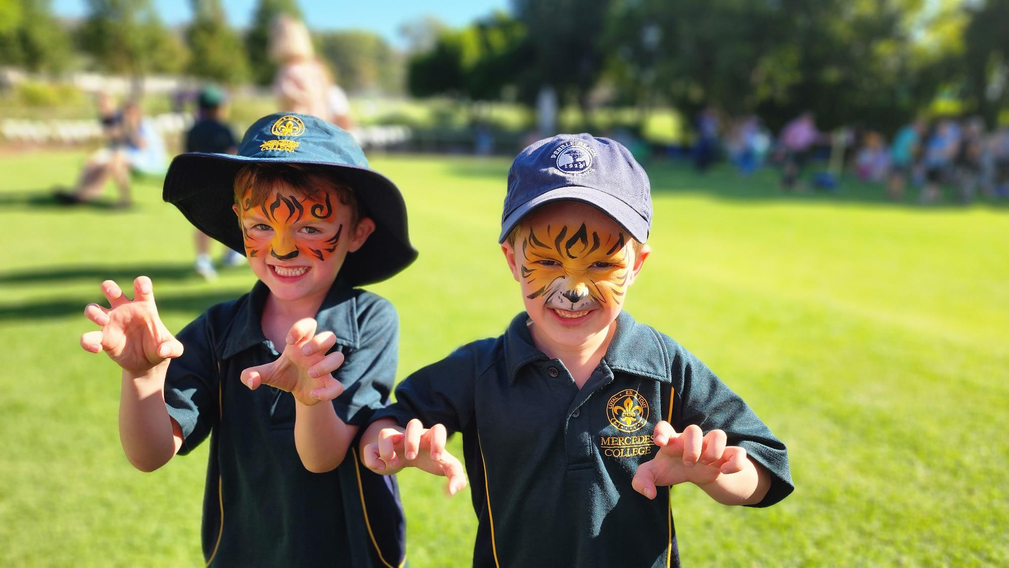 Two young boys with tiger face paint smiling and posing with claw hands on a grassy field at Mercedes College.