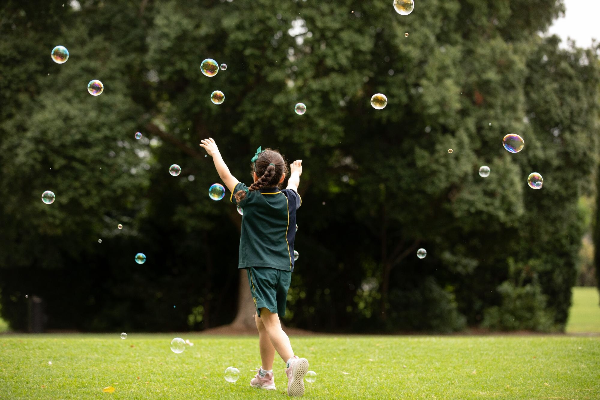 Mercedes College student in green uniform joyfully chasing bubbles across a grassy field, surrounded by trees.