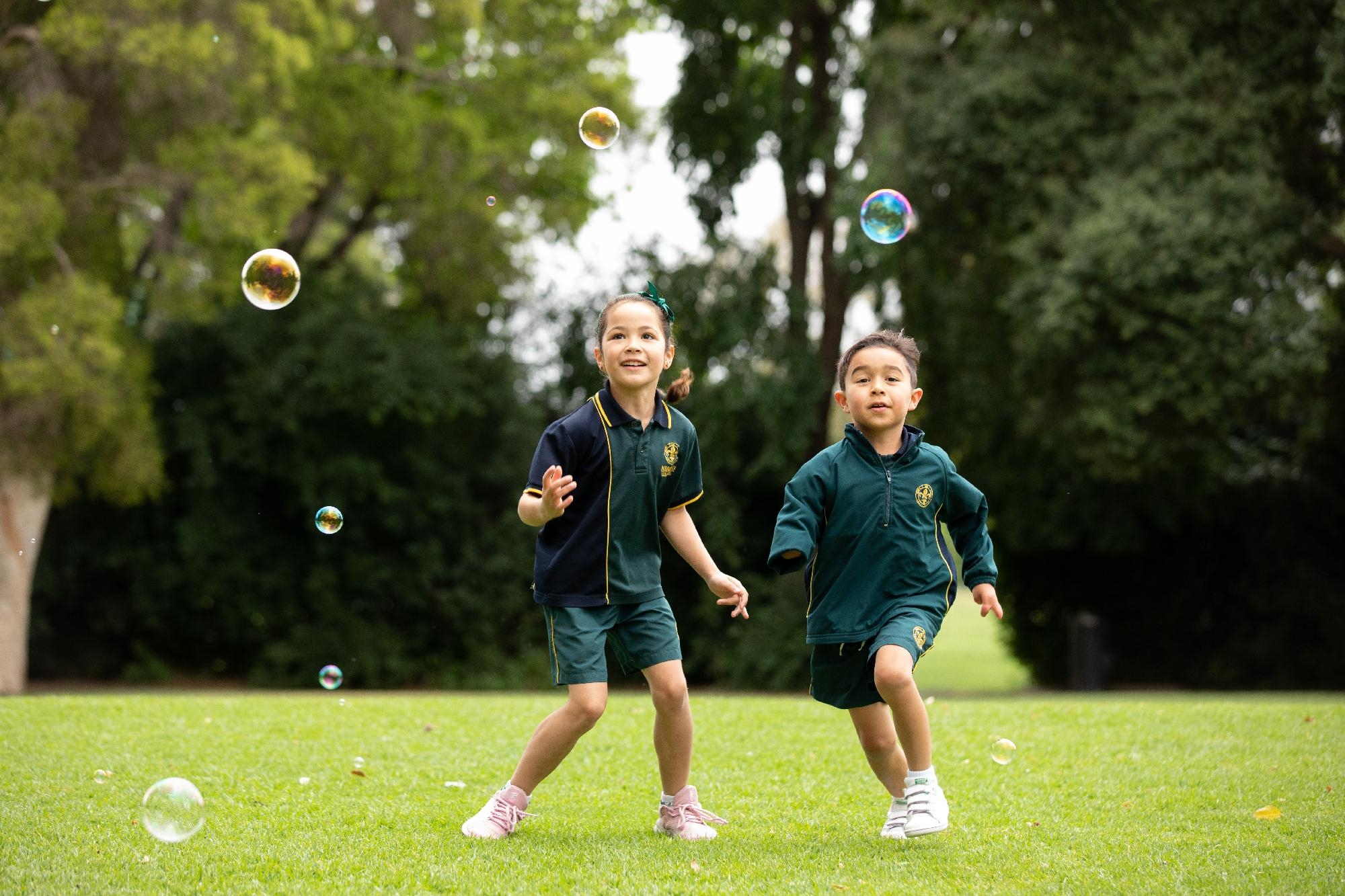 Young boy and girl in Mercedes College sports uniform chasing bubbles outdoors.