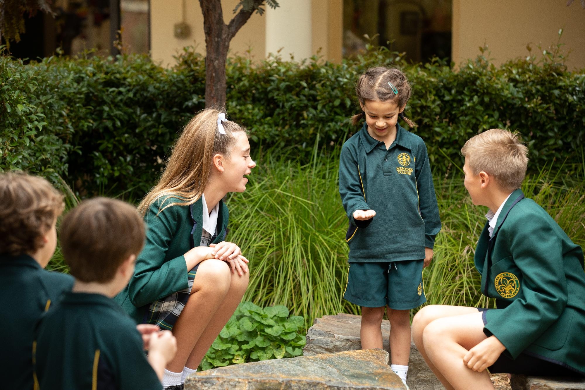 Mercedes College students gathered in a garden space, with a younger student showing something in her hand to smiling older students.