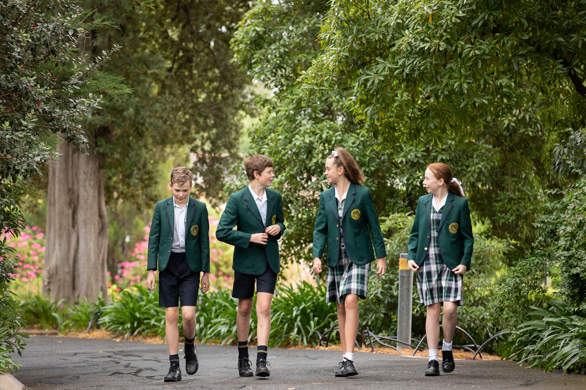 Four students in green school blazers walking together along a tree-lined path on campus.