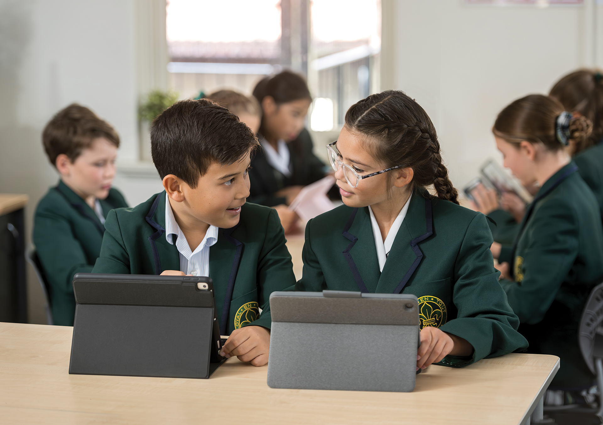 Two students in green school blazers working on tablets together in a classroom
