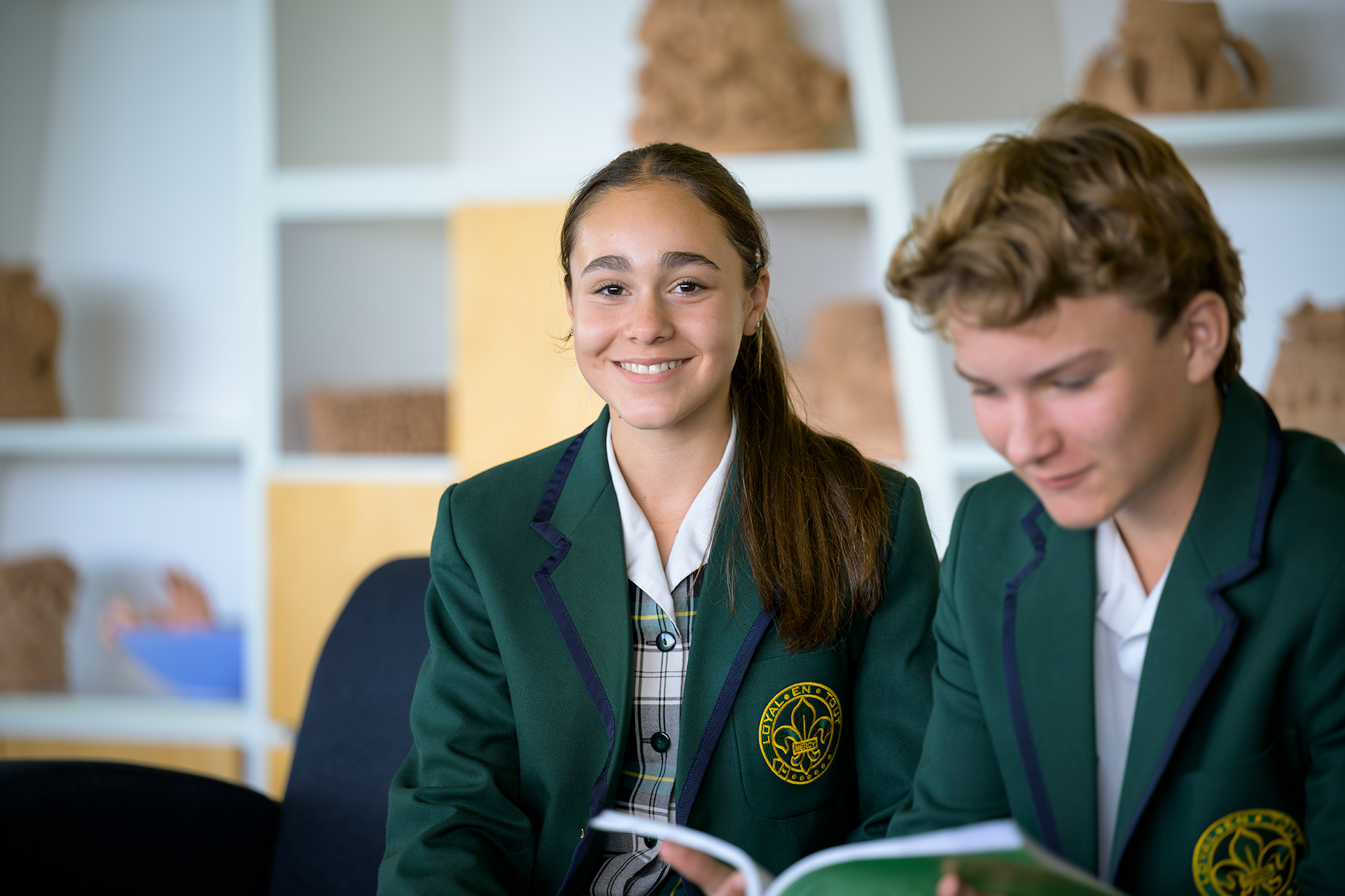Two Mercedes College students in green blazers reading together in a classroom with art displays in the background.