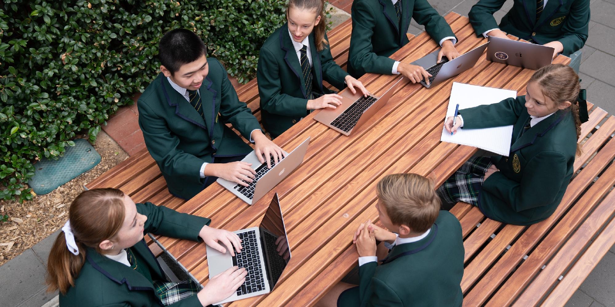 Mercedes College secondary students in formal green blazers collaborate on laptops and notebooks at an outdoor bench, engaging in a digital learning activity surrounded by greenery.
