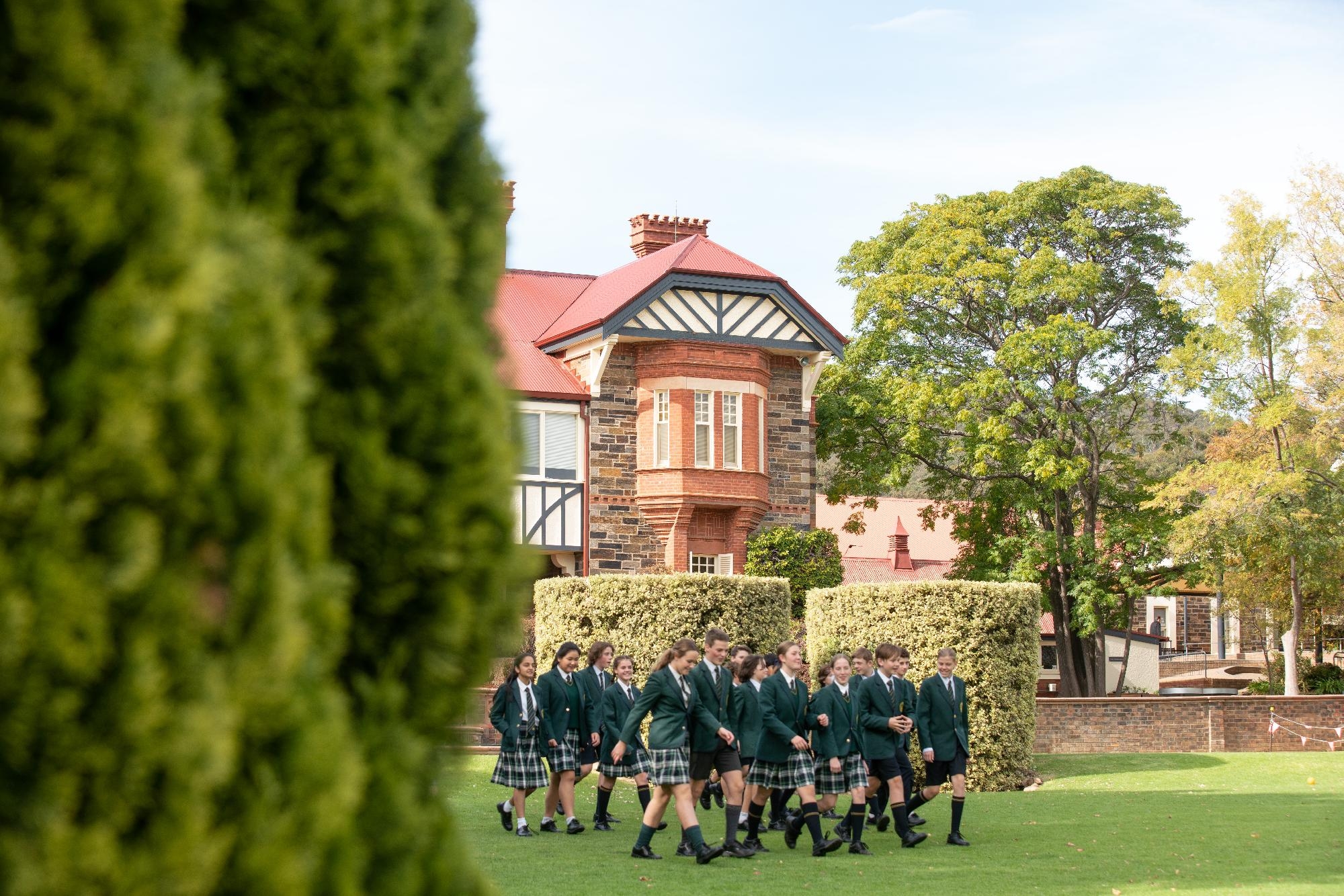 Mercedes College students in uniform walk in formation across the green lawn near a red-roofed stone building.