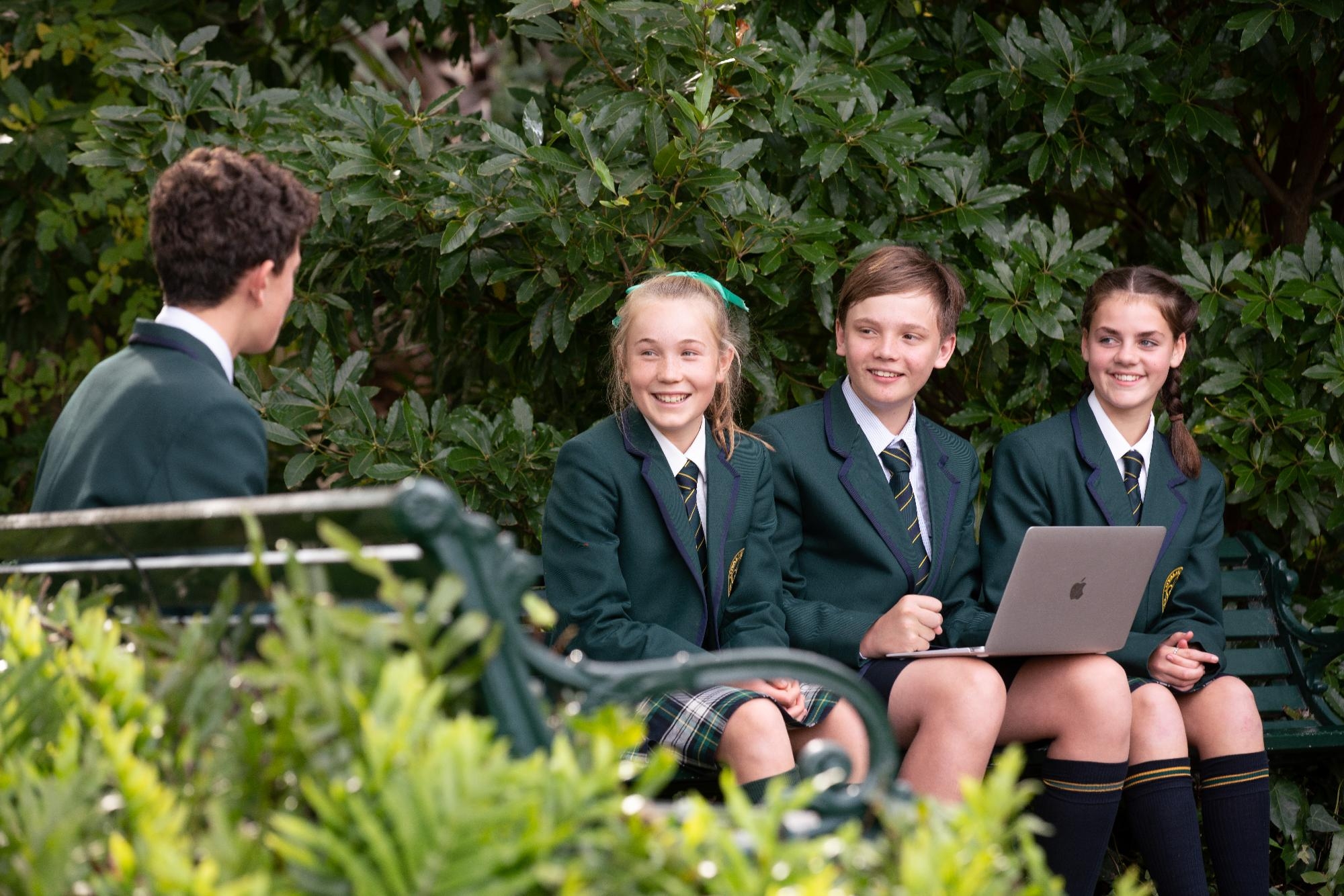 Smiling Mercedes College students in uniform using a laptop while enjoying the garden setting.