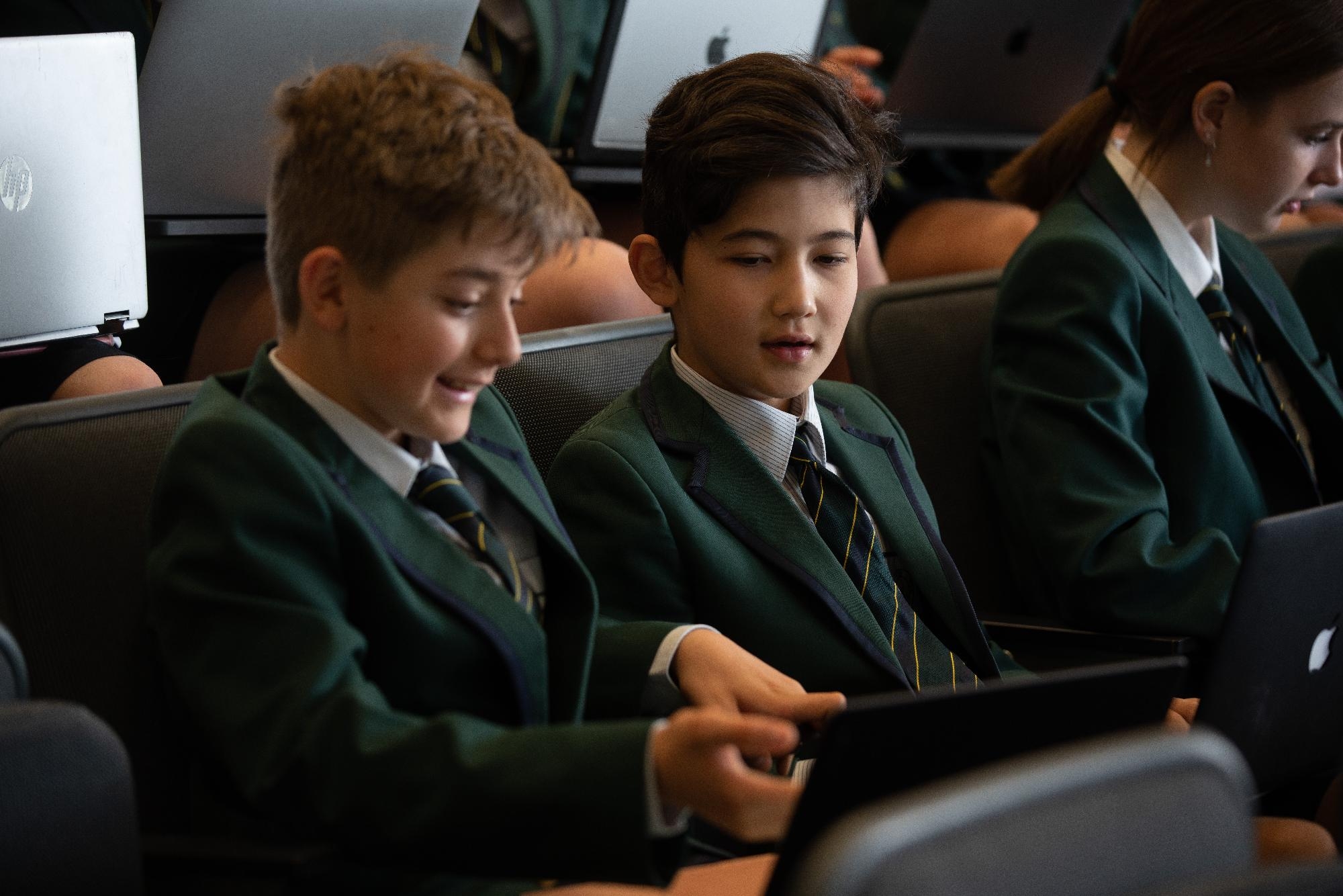 Two Mercedes College students use laptops side by side in a lecture setting, collaborating on their work.