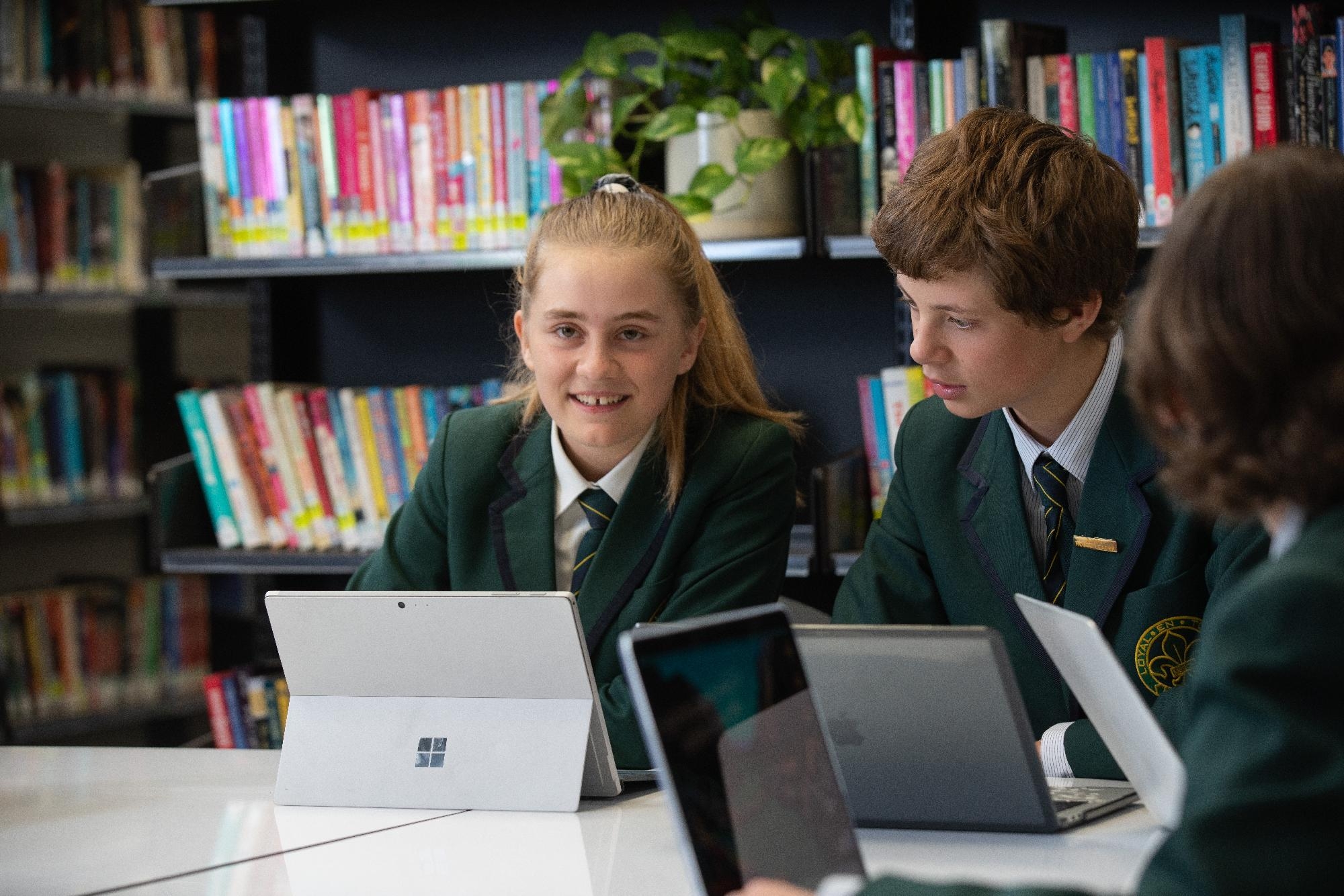A group of Mercedes College students in green blazers use digital devices together in the library, surrounded by bookshelves.