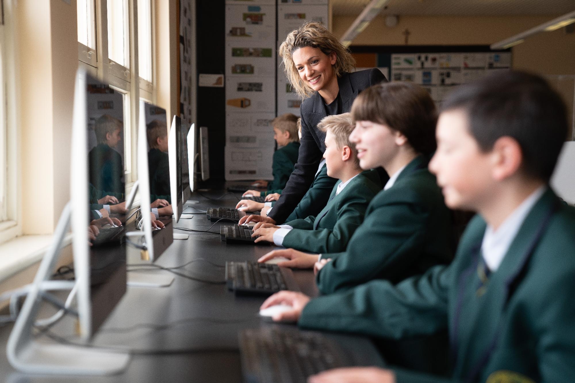 Mercedes College students in green blazers working at desktop computers in a modern classroom, guided by a smiling teacher.