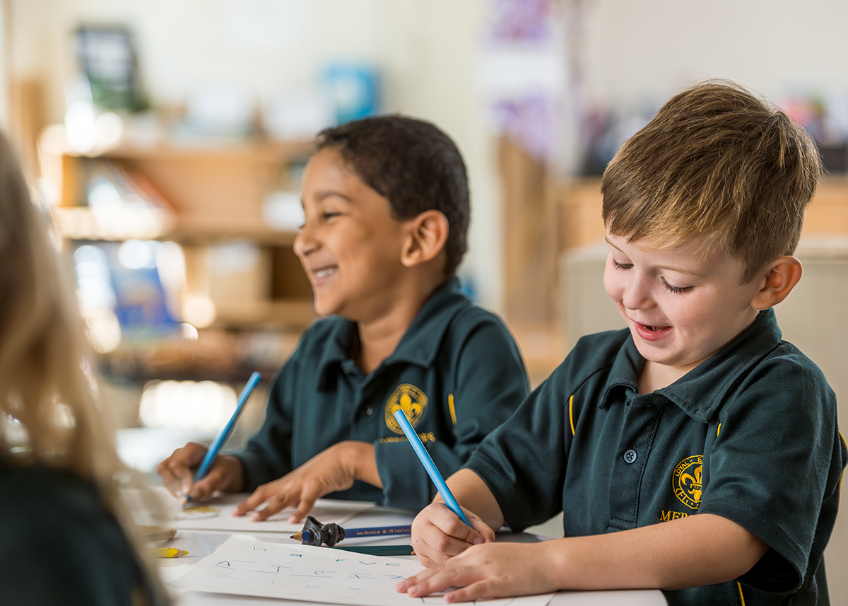 Two young students in school uniforms smiling and drawing with pencils at a classroom table.