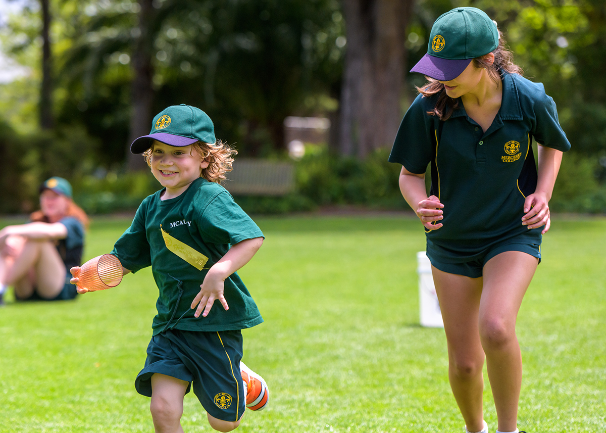 Smiling young student running on the grass with an older student beside them during outdoor school sports.