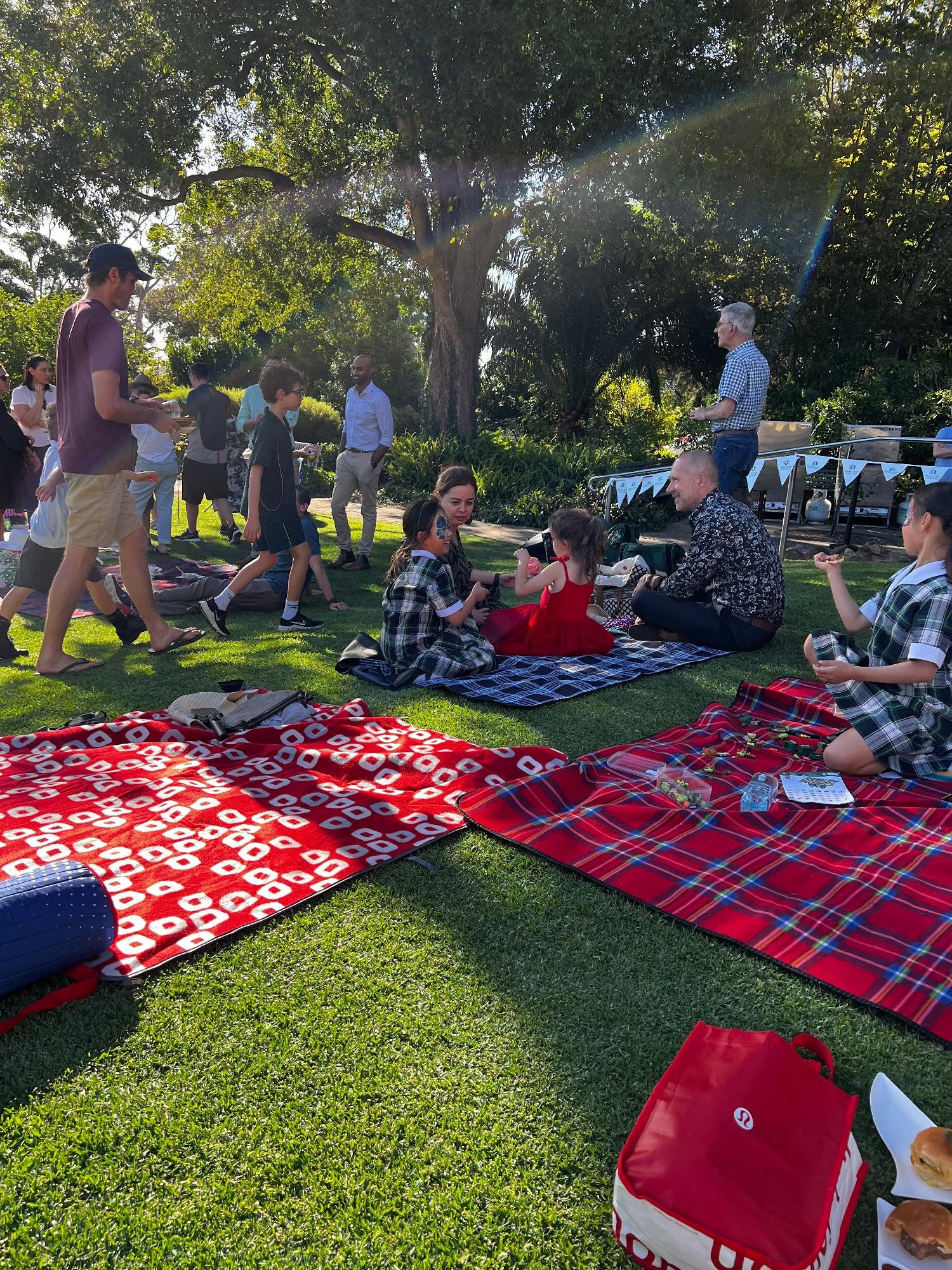 Mercedes College families enjoying a sunny afternoon picnic together on colourful blankets under the trees.