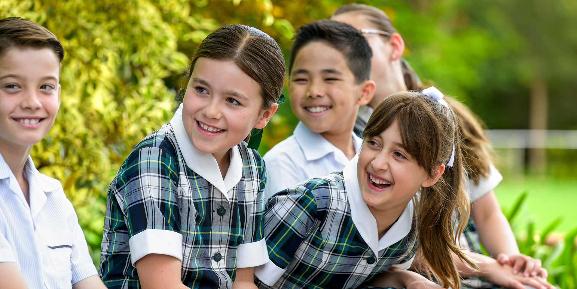 Mercedes College Junior School students smiling and sitting outdoors, wearing their green and navy checked uniforms on a bright, sunny day.