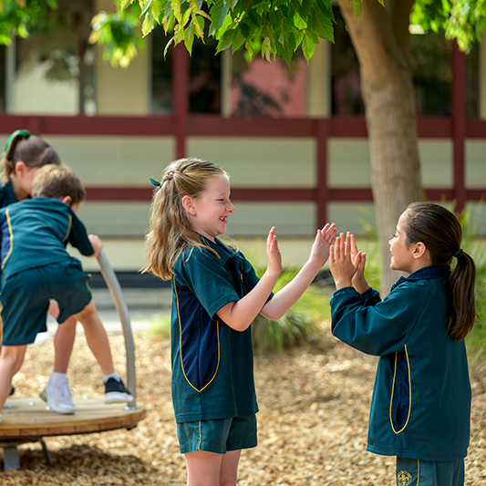 oung students at Mercedes College enjoying playtime outdoors, with two girls clapping hands and other children climbing on playground equipment in the background