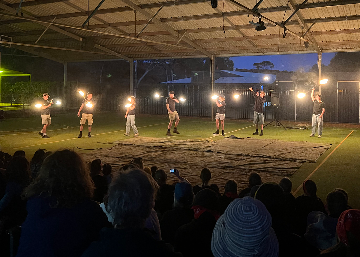 Students performing a fire dancing routine under a covered outdoor court, watched by an evening audience.