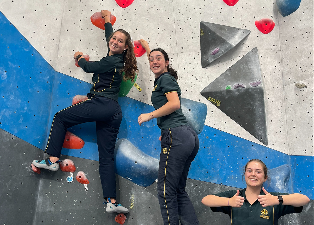 Two Mercedes College students climbing a rock wall while another student smiles with thumbs up.
