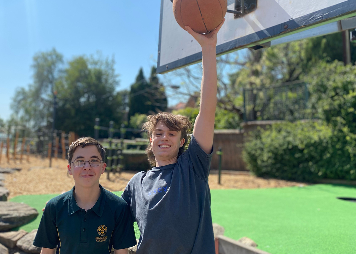 Two students smiling under a basketball hoop, with one playfully holding the ball above the ring on a sunny schoolyard.