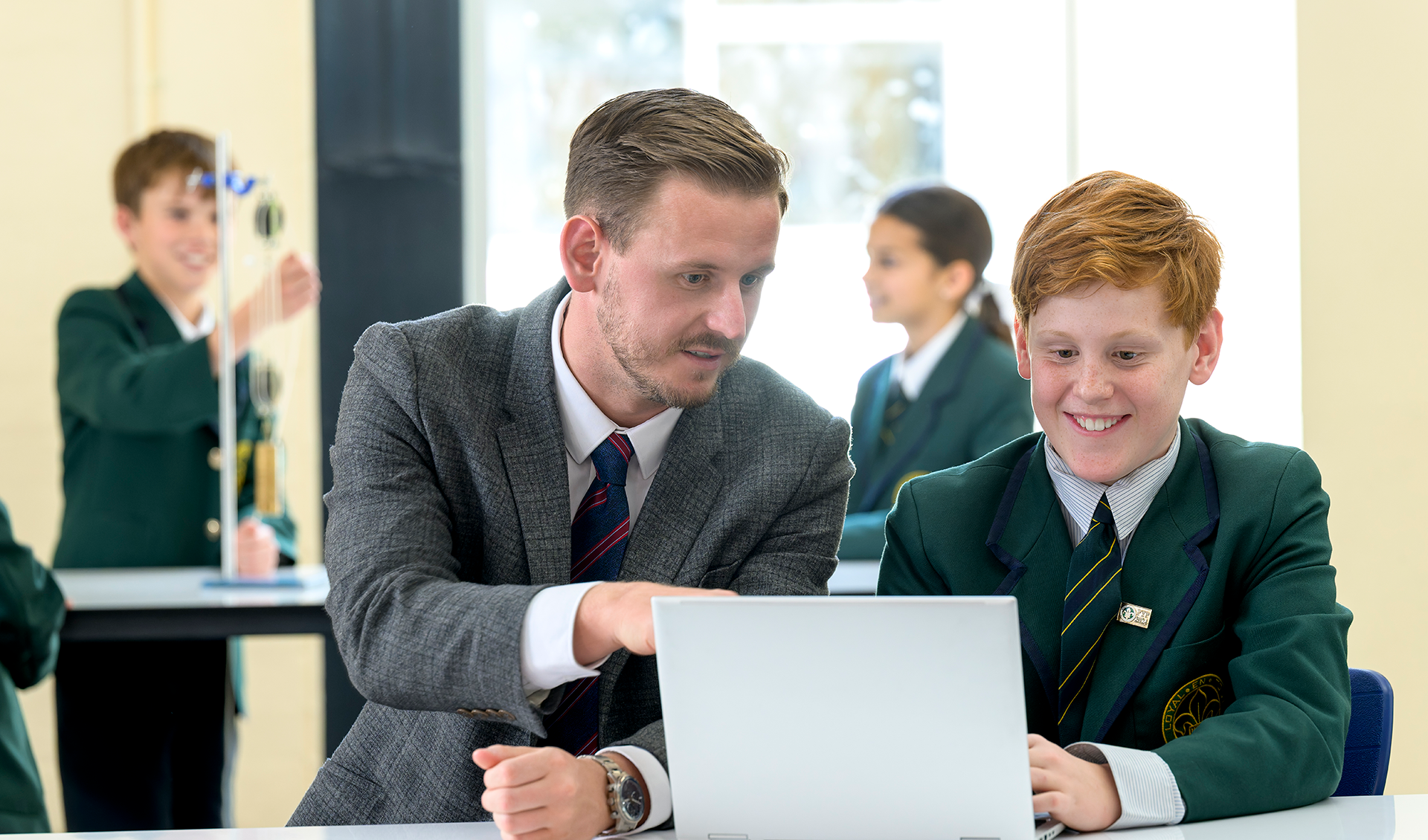 Teacher guiding a student on a laptop in a classroom with other students working in the background