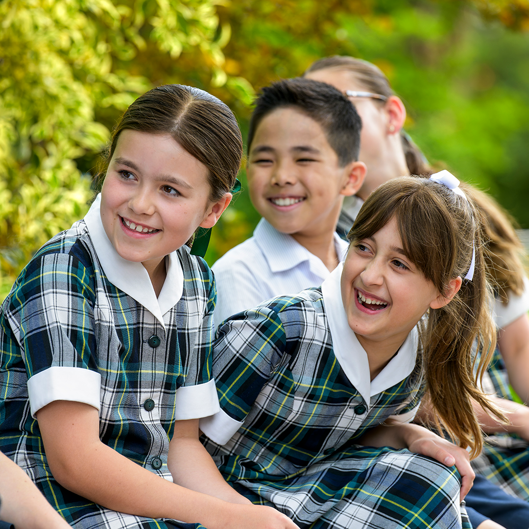 Mercedes College Junior School students smiling and laughing together outdoors, wearing green and navy checked uniforms.