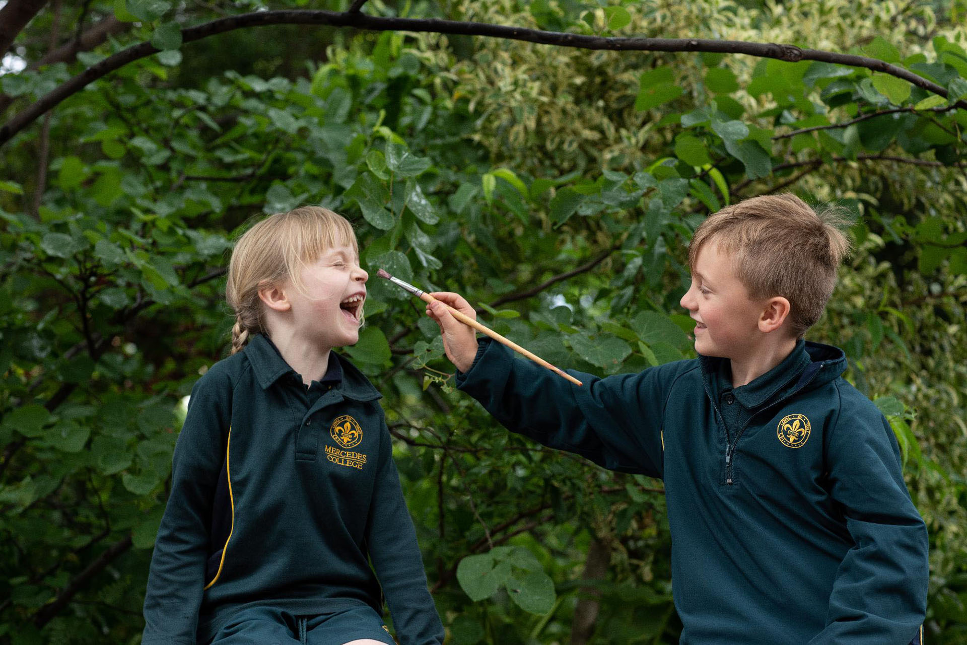 Young Mercedes College students dressed in green school uniforms, having fun as one paints the other's face during an outdoor activity.